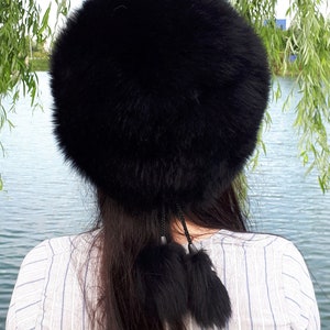 Peut inclure: Un chapeau rond en fausse fourrure noire et duveteuse, agr&eacute;ment&eacute; de deux pompons en fourrure noire suspendus par de fines cordelettes tress&eacute;es. Le chapeau pr&eacute;sente un aspect dense et textur&eacute; avec une &eacute;paisseur de fibres noires volumineuses.