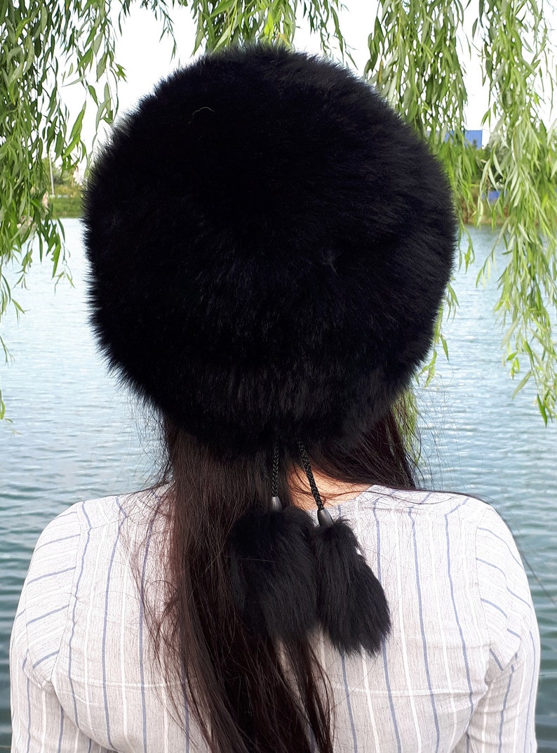 Peut inclure: Un chapeau rond en fausse fourrure noire et duveteuse, agr&eacute;ment&eacute; de deux pompons en fourrure noire suspendus par de fines cordelettes tress&eacute;es. Le chapeau pr&eacute;sente un aspect dense et textur&eacute; avec une &eacute;paisseur de fibres noires volumineuses.