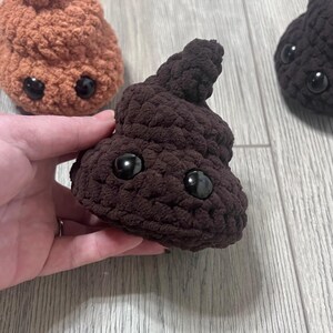 Puede incluir: Emoji de caca marrón tejido con grandes ojos negros. El emoji se sostiene en una mano, con otros dos emojis similares en el fondo, uno en marrón claro y otro en marrón oscuro.
