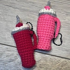 May include: Two crocheted milkshake keychain charms, one pink and one red, with white whipped cream and a gray straw on top. Each charm has a black chain attached.