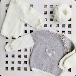 Könnte beinhalten: Zwei gestrickte Babysweater, einer weiß und einer grau mit weißen Akzenten. Der graue Pullover hat ein weißes Tiergesichtsdesign und Knöpfe.