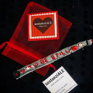 May include: A gray denim bracelet with red stitching and red beads. The bracelet has the words "Be Still" stitched on it. The bracelet is packaged in a red organza bag with a red and white card that says "sostanziALE Anelli & Co".