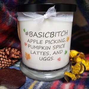 Puede incluir: Una vela blanca con tapa negra y lazo blanco. La vela tiene el texto "#BASICBITCH APPLE PICKING, PUMPKIN SPICE LATTES, AND UGGS". La vela está sobre una base de vidrio transparente.