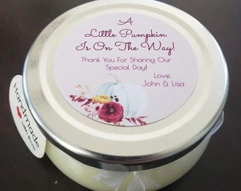 May include: A small, round jar with a silver lid and a cream-colored body. The lid features a label with the text "A Little Pumpkin Is On The Way!" and a floral and pumpkin design. A small tag reads "Handmade with love."