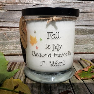 Peut inclure: Une bougie blanche dans un bocal en verre avec un couvercle noir. La bougie porte l'inscription "Fall Is My Second Favorite F-Word" avec des graphiques de feuilles d'automne. Une petite étiquette indique "Handmade with love."