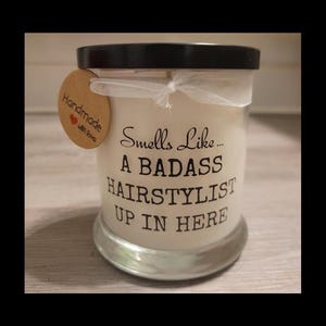 Puede incluir: Una vela blanca en un tarro de cristal transparente con tapa negra. La vela tiene el texto "Smells Like... A BADASS HAIRSTYLIST UP IN HERE" impreso. Una pequeña etiqueta con "Handmade with love" está adjunta.