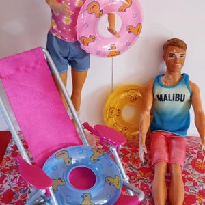 May include: A beach scene with two dolls, one holding a pink inflatable ring with duck designs. The other doll wears a blue tank top with "MALIBU" printed on it. A pink beach chair and several inflatable rings are also present.