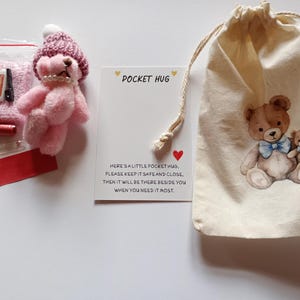 May include: A pink teddy bear with a knitted hat and pearl necklace, alongside a small bag of pink beads, a red tag, and a drawstring bag with two teddy bear illustrations. The card reads "POCKET HUG".