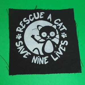 Puede incluir: Parche de tela negro con texto blanco y un gato de dibujos animados. El texto dice "Rescata un gato, salva nueve vidas".