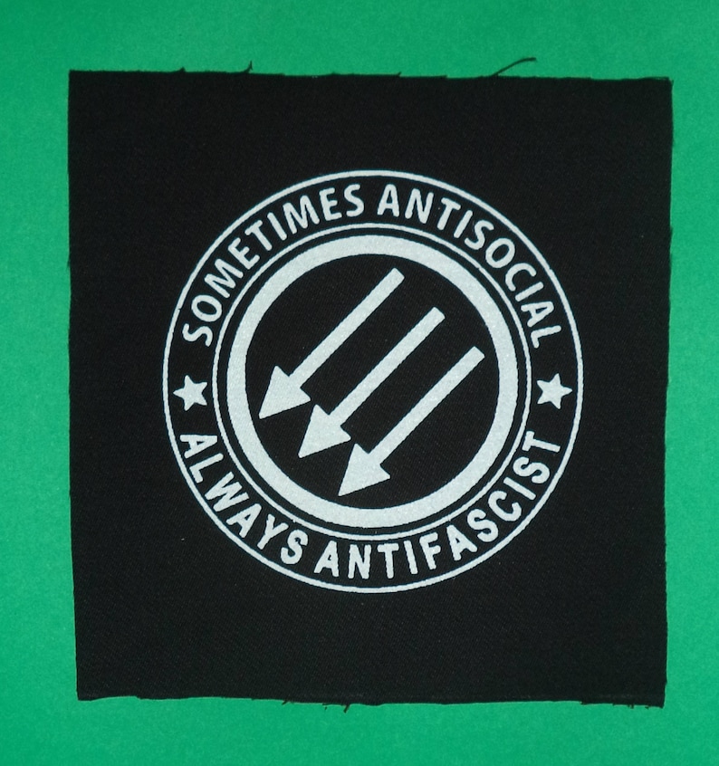 May include: Black and white patch with a circle containing three white arrows pointing to the left. The circle has the text "Sometimes Antisocial Always Anti-Fascist" around it.