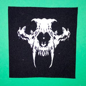 Puede incluir: Parche de tela negro con una calavera blanca bordada con dientes grandes.