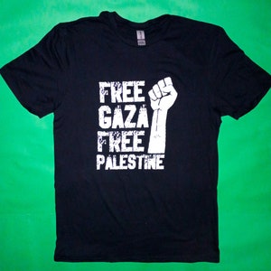 Può includere: Maglietta nera con testo bianco che recita "FREE GAZA FREE PALESTINE" e un grafico di un pugno alzato.
