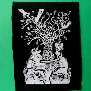 May include: Black and white embroidered patch featuring a detailed illustration of a human head with a tree growing out of it. The tree has many eyes and branches with hands reaching out.
