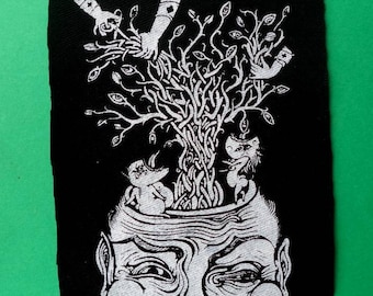 May include: Black and white embroidered patch with a detailed illustration of a human head with a tree growing out of it. The tree has many eyes and branches with hands reaching out.