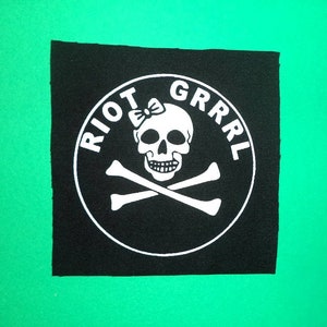 Pode incluir: Remendo de tecido preto com um círculo branco contendo uma caveira com um laço, ossos cruzados e o texto "RIOT GRRRL".