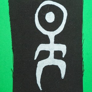 Könnte beinhalten: Eine weiße stilisierte Figur mit einem Kreis als Kopf und einem kleinen Kreis im Kopf auf einem schwarzen Stoffhintergrund.