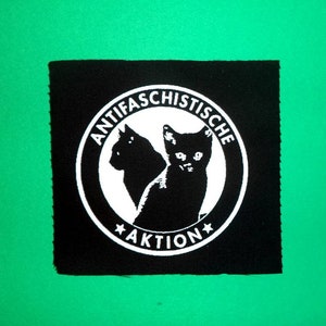 Könnte beinhalten: Schwarz-weißes Patch mit einem Kreis, der den Text "ANTIFASCHISTISCHE AKTION" und zwei schwarze Katzen enthält, die sich gegenüberstehen.
