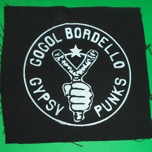 Pode incluir: Etiqueta bordada preta e branca com um design circular. O círculo apresenta uma estilingue com uma estrela acima e o texto "Gogol Bordello Gypsy Punks".