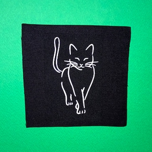 May include: A black fabric patch with a white outline of a cat walking. The cat is facing forward with its tail curled around its body.