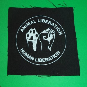 Peut inclure: Écusson en tissu noir avec du texte et des images blancs. Le texte dit "Animal Liberation Human Liberation". Les images sont une empreinte de patte et un poing.
