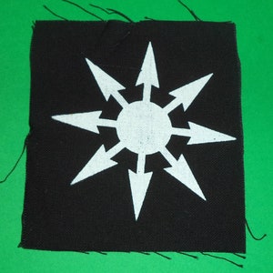 Peut inclure: Écusson en tissu noir avec une étoile blanche à huit branches avec des flèches pointant vers l'extérieur. L'étoile a un cercle blanc au centre.