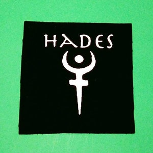 May include: Black patch with white text that reads "HADES" and a white symbol of a crescent moon with a circle in the center and a dagger below.