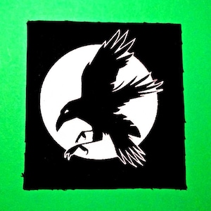 Może przedstawiać: Czarno-biała łatka z sylwetką kruka lecącego przed białym kołem. Łatka jest przyszyta do zielonego tła.