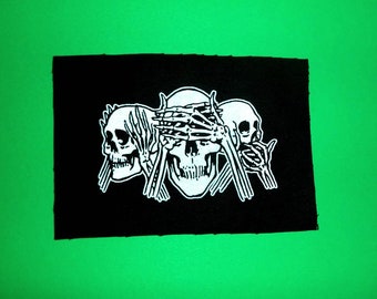 May include: Black and white embroidered patch featuring three skulls with hands covering their eyes. The patch is on a black background.