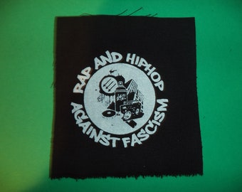 May include: Black fabric patch with white text and graphic. The text reads "Rap and Hip Hop Against Fascism". The graphic is a spray can, a record, a boombox, and a microphone.