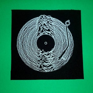 Könnte beinhalten: Ein schwarzer Stoff-Patch mit einer weißen Umrandung einer Schallplatte auf einem Plattenspieler. Die Schallplatte hat ein Wellenmuster und der Plattenspieler hat einen Tonarm.