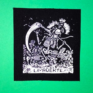 Pode incluir: Patch bordado preto e branco com um esqueleto segurando uma foice com o texto "LA MUERTE" na parte inferior.