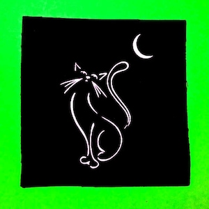 Op de afbeelding: Een zwarte stoffen patch met een wit silhouet van een kat die naar een halve maan kijkt.