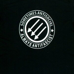 May include: Black t-shirt with a white circle graphic that says "Sometimes Antisocial Always Anti-Fascist" and features three white arrows pointing to the left.