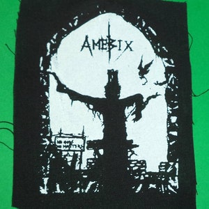 Op de afbeelding: Zwart-witte geborduurde patch met de bandnaam "Amebix" in een gotisch lettertype. De patch toont een silhouet van een persoon met uitgestrekte armen voor een grote boog.