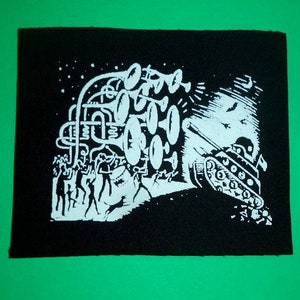 Könnte beinhalten: Schwarz-weiß gestickter Patch, der einen Panzer zeigt, der von einer Gruppe von Menschen, die Trompeten spielen, nach vorne geschoben wird. Der Patch hat einen abgenutzten Look und ist auf einen schwarzen Stoff genäht.