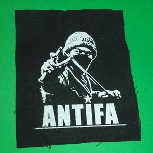Può includere: Toppa nera con una grafica bianca di una persona con berretto e passamontagna, che usa una fionda. La parola "ANTIFA" è stampata in lettere bianche sotto.