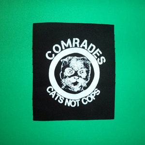 Peut inclure: Patch noir et blanc avec un cercle contenant un visage de chat et le texte "Comrades Cats Not Cops".