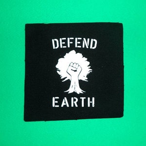 Puede incluir: Parche cuadrado negro con texto y gráfico en blanco. El texto dice "DEFEND EARTH" con un gráfico de árbol que tiene un puño en el centro. El parche está sobre un fondo verde.