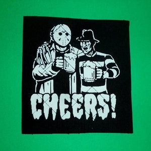 Peut inclure: Patch noir et blanc avec des illustrations de Jason Voorhees et Freddy Krueger tenant des bières. Le texte "CHEERS!" est sous les personnages.