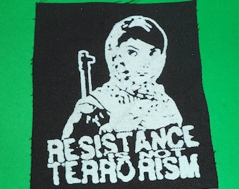 May include: Black and white embroidered patch with a person wearing a mask and holding a rifle. The text "RESISTANCE IS NOT TERRORISM" is below the image.