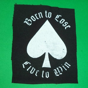 Peut inclure: Écusson noir avec texte blanc et symbole de pique blanc. Le texte indique "Born to Lose" au-dessus de la pique et "Live to Win" en dessous. L'écusson est rectangulaire avec un bord légèrement effiloché.