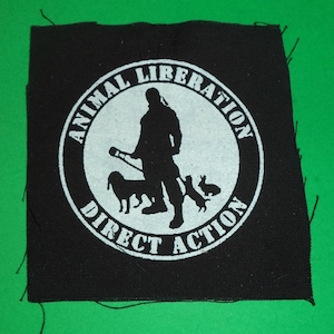 May include: Black and white patch with the text "Animal Liberation Direct Action" in a circle. A silhouette of a person holding a stick with two animals beside them is in the centre of the circle.