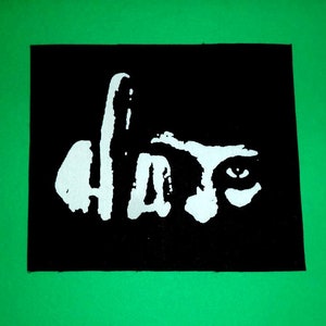 May include: A black square patch featuring the word "Hate" in white, designed to look like a face. The design includes an eye and a partial profile. The patch is set against a bright green background.