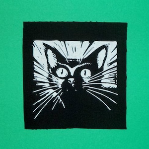 Könnte beinhalten: Ein schwarz-weiß Linolschnitt eines Katzenkopfes mit großen Augen und Schnurrhaaren. Die Katze ist vor einem schwarzen Quadrat vor einem grünen Hintergrund platziert. Die Merkmale der Katze sind mit kräftigen Linien stilisiert.