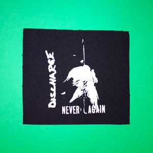 Peut inclure: Patch de groupe Discharge noir et blanc avec le texte "Never Again" et le nom du groupe "Discharge" écrit verticalement.