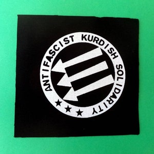 Peut inclure: Patch noir et blanc avec le texte "Anti-fasciste Solidarité Kurde" dans un cercle avec trois flèches blanches pointant vers la droite.