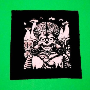 Könnte beinhalten: Schwarz-weiß-Patch mit einer Grafik eines Außerirdischen mit einem schädelartigen Gesicht und einem großen Kopf. Der Außerirdische ist von fliegenden Untertassen umgeben. Der Patch befindet sich auf einem grünen Hintergrund.