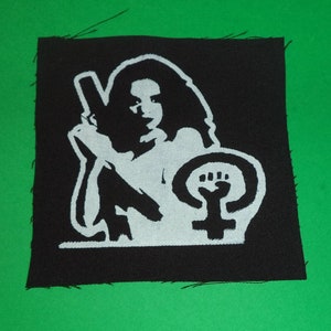 Könnte beinhalten: Schwarz-weiß gestickter Aufnäher mit einer Frau, die eine Waffe hält, und einem feministischen Symbol mit einer Faust.