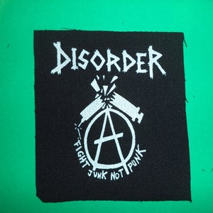 Peut inclure: Patch brodé noir et blanc avec le texte "DISORDER" dans une police de style punk rock. Le patch présente un cercle avec un symbole d'anarchie et deux seringues qui se croisent. Le texte "FIGHT JUNK NOT PUNK" est écrit autour du cercle.