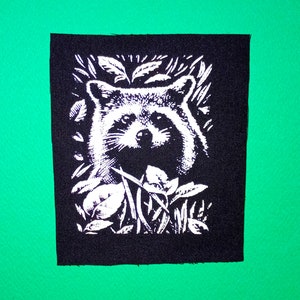 以下が含まれることがあります： 葉と枝に囲まれたアライグマの顔の白黒刺繍パッチ